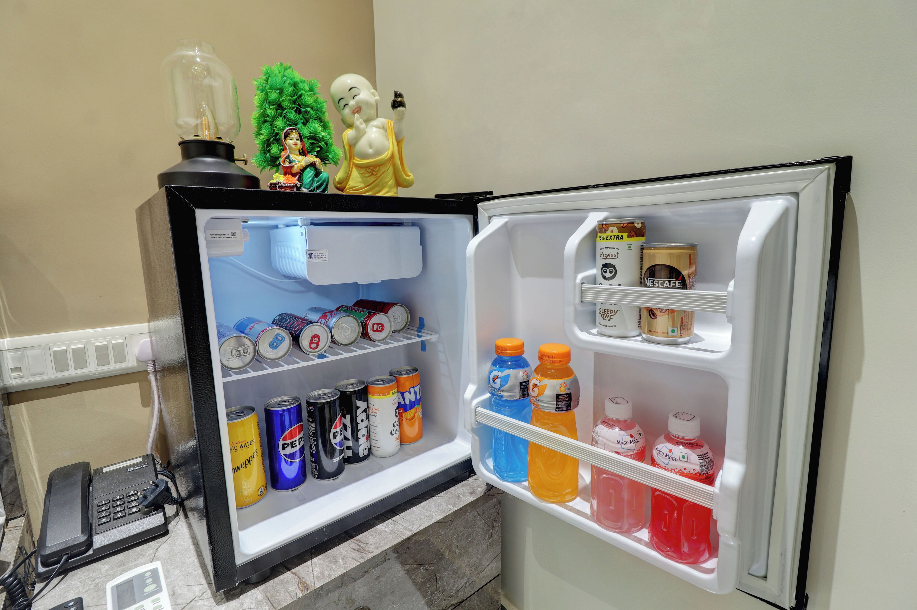 Un refrigerador lleno de bebidas y aperitivos, un teléfono y una pequeña estatua de Buda encima.