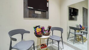 A room with two grey chairs, a small round table with snacks, a wall-mounted TV, and a large mirror reflecting the room. - Royal Inn (Navi Mumbai)