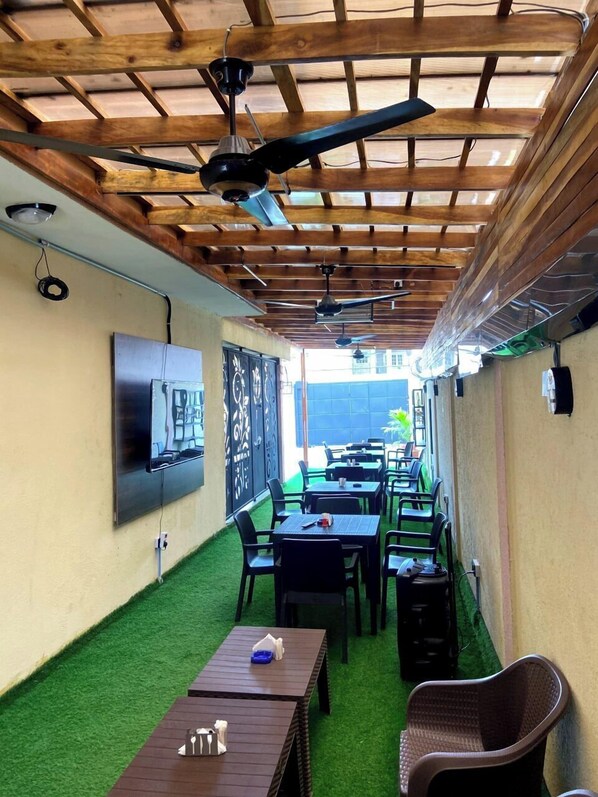 A covered outdoor seating area with tables and chairs, a ceiling fan, and a TV mounted on the wall.