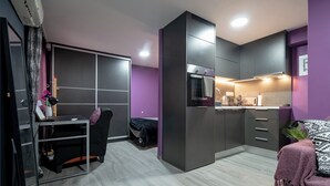 Um apartamento estúdio compacto com cozinha compacta, uma cama com cobertor roxo e uma mesa com computador.