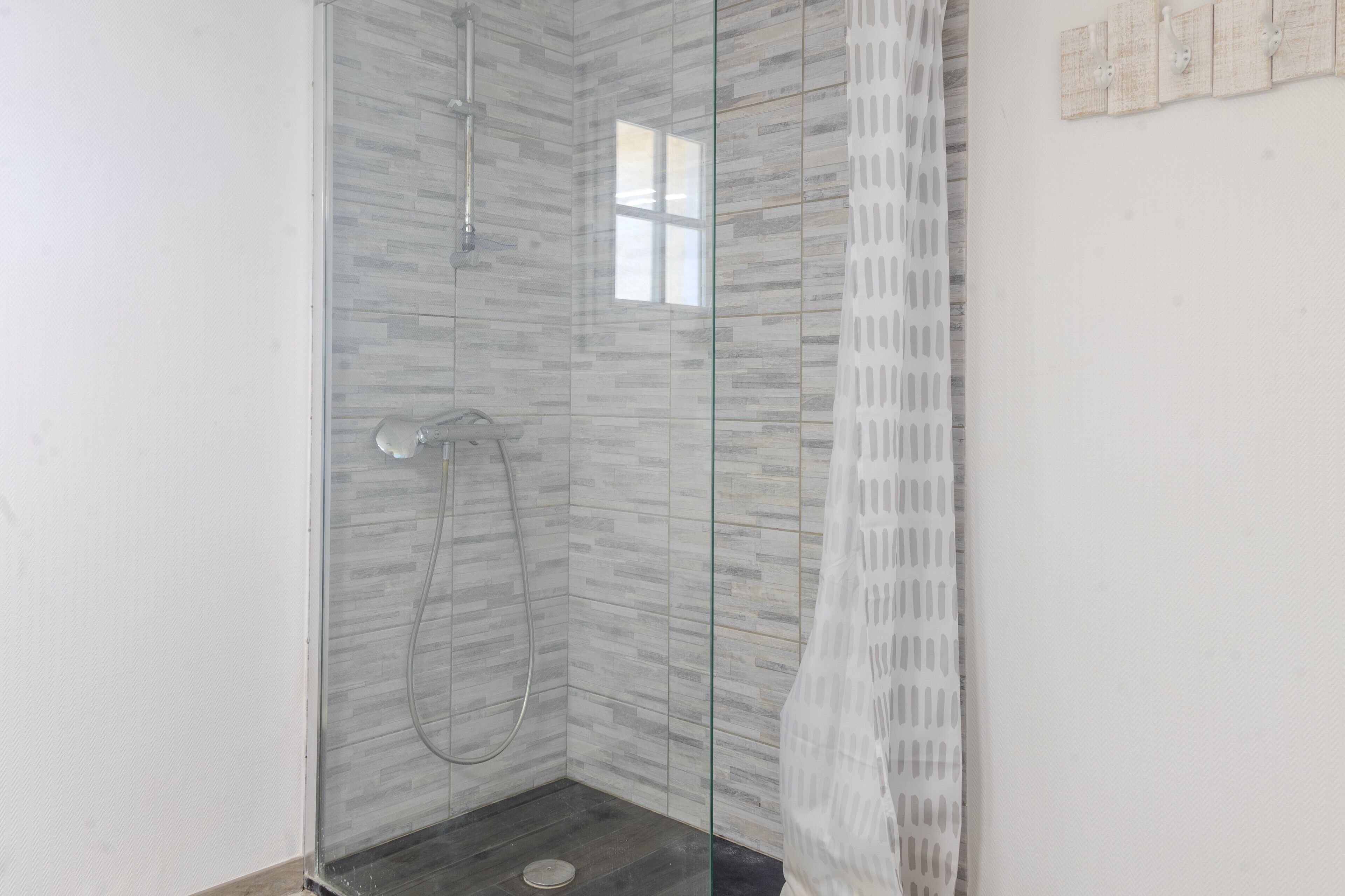 Eine gläserne Dusche mit gefliesten Wänden, einem kleinen Fenster und einem weißen Vorhang.