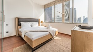 A modern hotel room with a wooden bed, a bedside table with a lamp, a large window with a city view, and a small desk with a bowl.