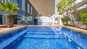 A swimming pool with blue mosaic tiles, surrounded by a modern building with white and grey walls, and a palm tree.