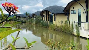 A row of modern, single-story buildings with flat roofs, situated beside a calm water body, surrounded by lush greenery. - Capybara Zen Resort And Spa (Mae Pong)