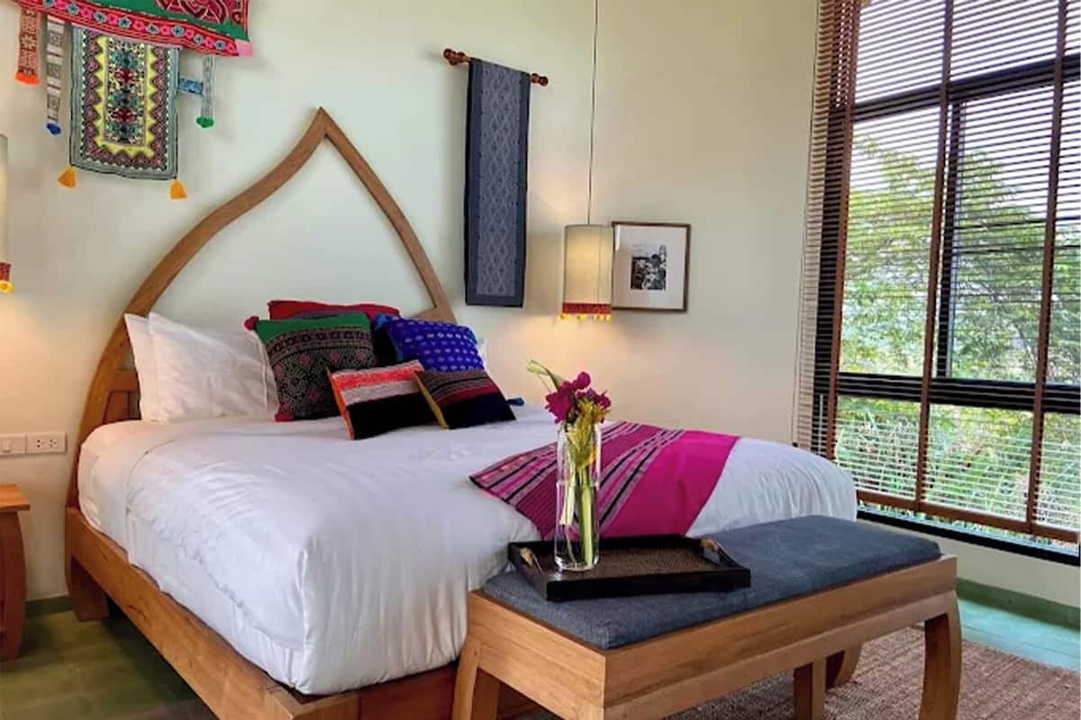 A bedroom with a wooden bed, colorful pillows, a vase with flowers, and a window with blinds.