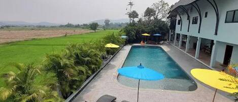 A swimming pool with lounge chairs and umbrellas, surrounded by greenery and a building with a curved roof.