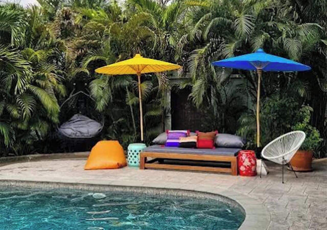 Ein Poolbereich mit einer Holzbank, bunten Kissen und zwei Sonnenschirmen.