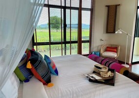 Un dormitorio con una cama grande, almohadones coloridos, una silla de mimbre y vistas al exterior.