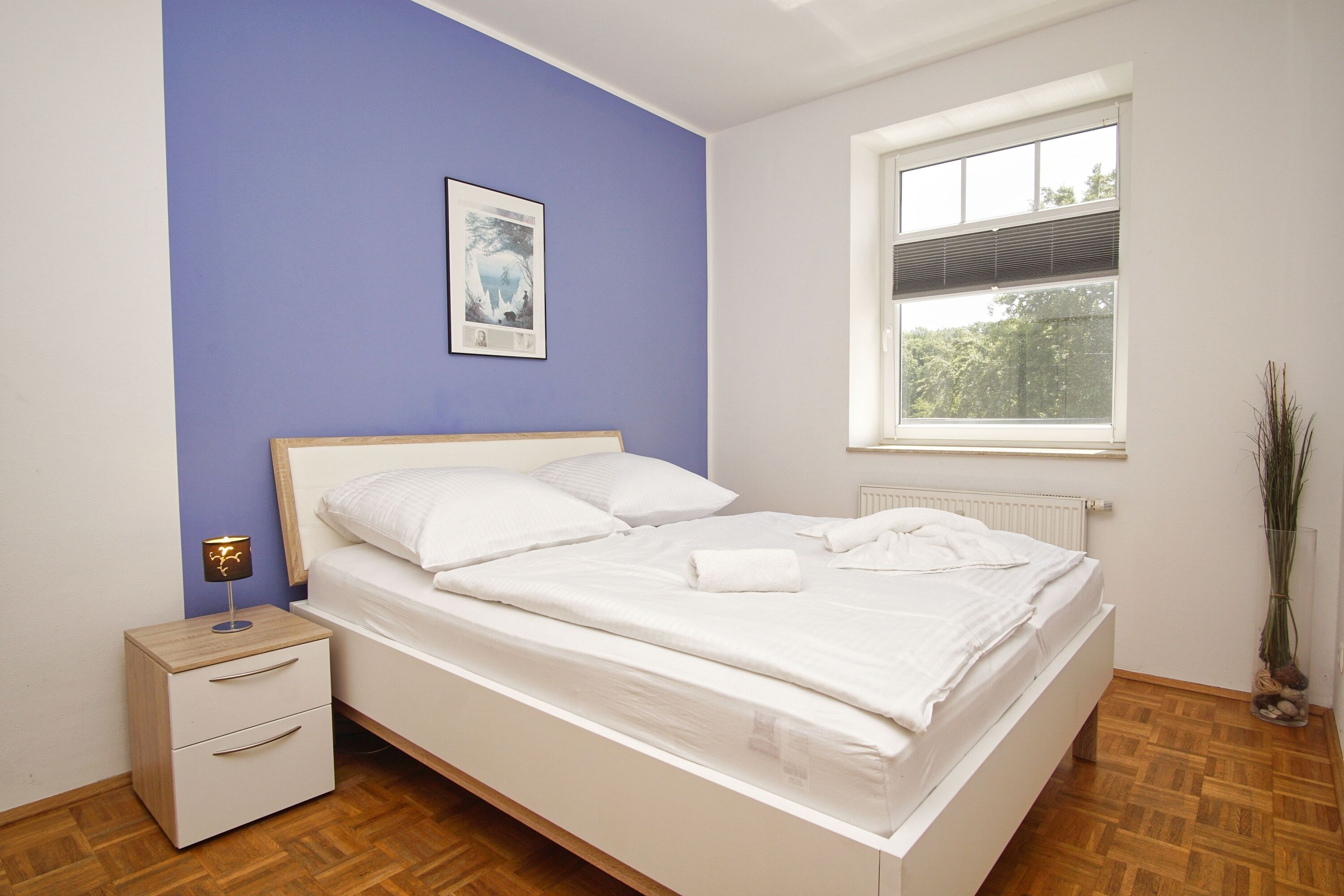 A neatly made bed in a room with a blue accent wall, a small bedside table with a lamp, and a window with a view of trees.