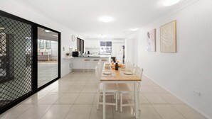 A modern kitchen with white cabinets, a dining area with a wooden table and white chairs, and a sliding glass door leading to an outdoor area. - Beach Break: Firepit & Outdoor Entertaining (Caloundra Region)