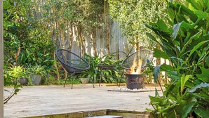 An outdoor patio with a wooden deck, two black metal chairs, a small table, and a fire pit. - Beach Break: Firepit & Outdoor Entertaining (Caloundra Region)