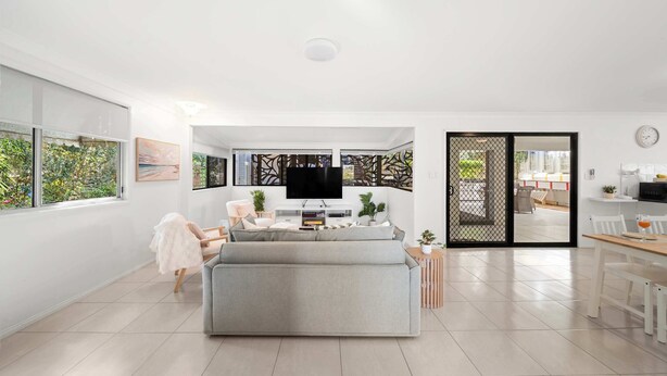 A modern living room with a grey sofa, a flat-screen TV, and a dining area with a wooden table and chairs. - Beach Break: Firepit & Outdoor Entertaining (Caloundra Region)