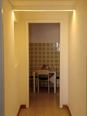 Un estrecho pasillo que conduce a un comedor con una mesa y sillas, un salpicadero de azulejos y una lámpara en el techo.