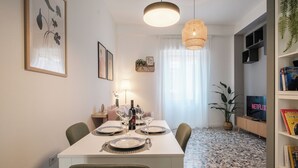 Et spiseområde med et hvidt bord dækket til fire personer, en hængende lampe og en reol med bøger samt et fjernsyn der viser 'Netflix'.