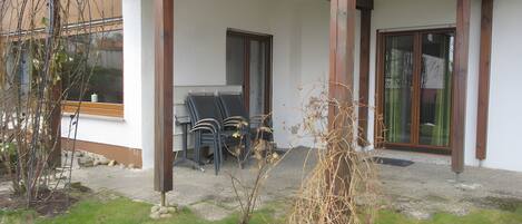 A covered outdoor seating area with chairs and a table, a wooden pergola, and a small garden with a plant and rocks.