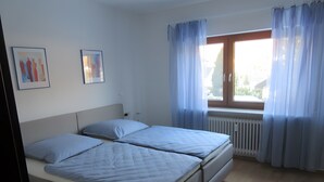 Een slaapkamer met twee bedden, een raam met gordijnen, een radiator en twee ingekaderde foto’s aan de muur.