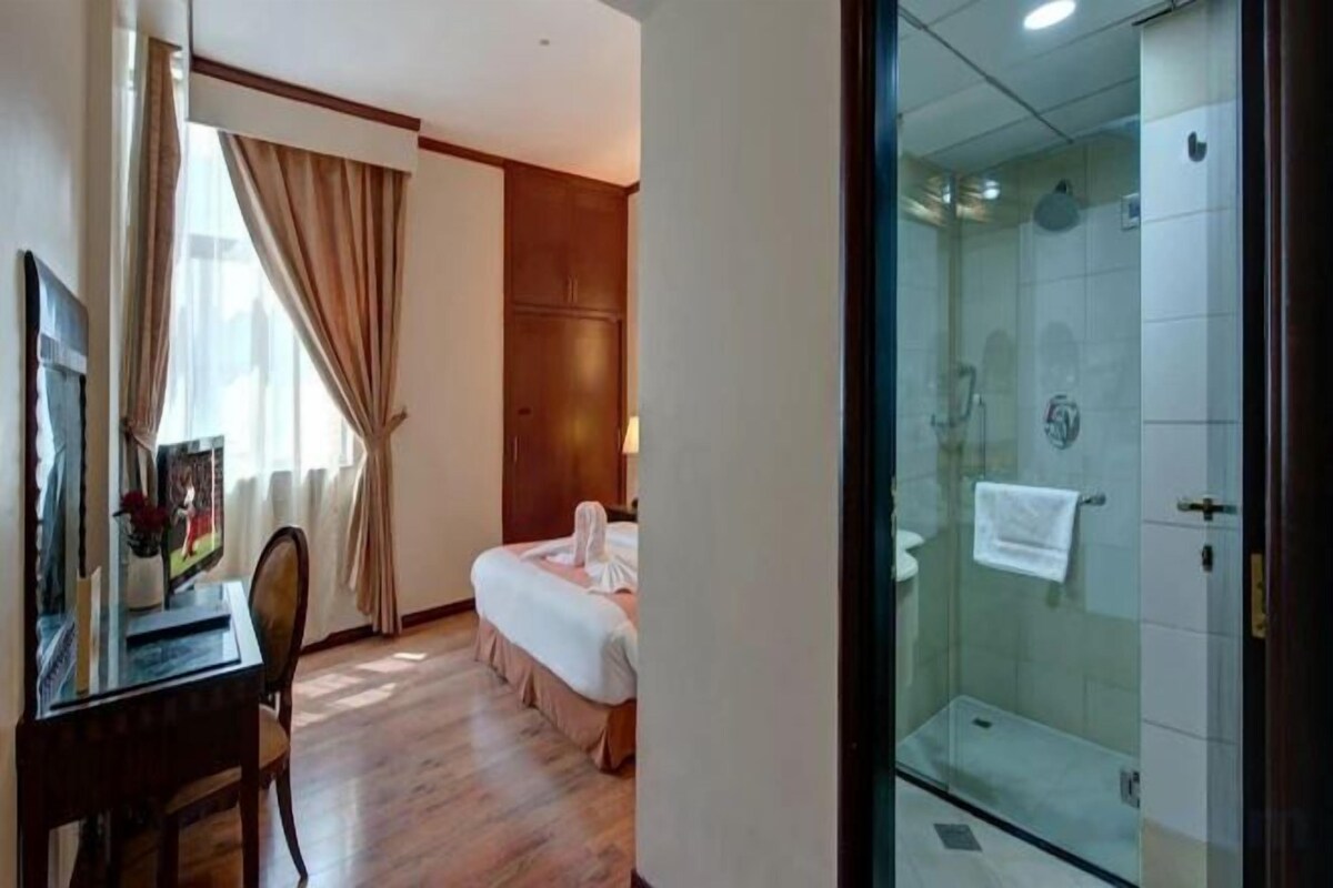 A hotel room with a bed, a desk, a chair, a shower area, and a glass door.