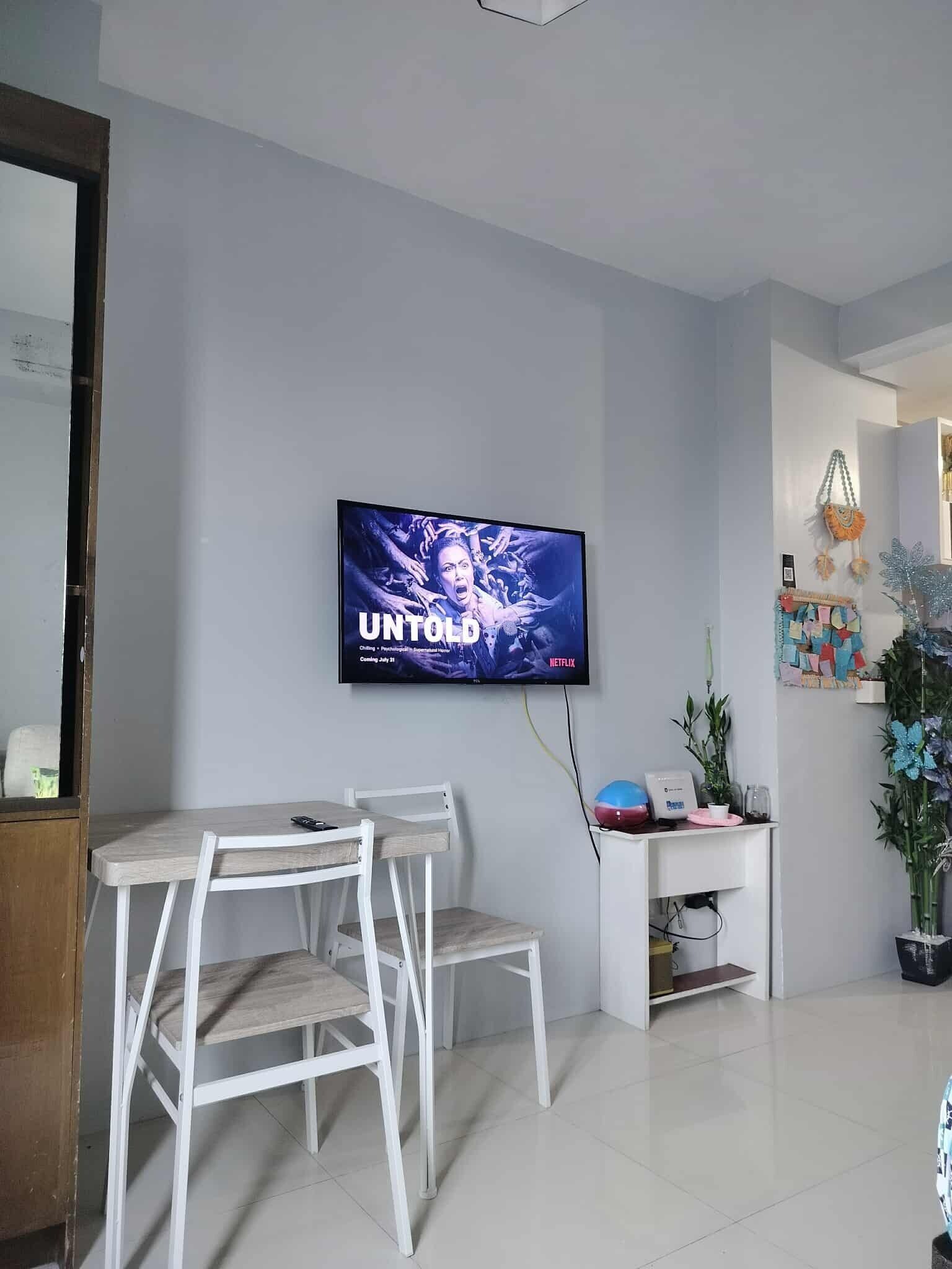 A modern living room with a flat-screen TV mounted on the wall, a white dining table with chairs, and a small white cabinet with decorative items.