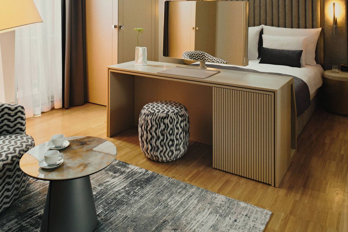 A modern hotel room with a skylight, a bed, a desk, and a seating area with a patterned ottoman.