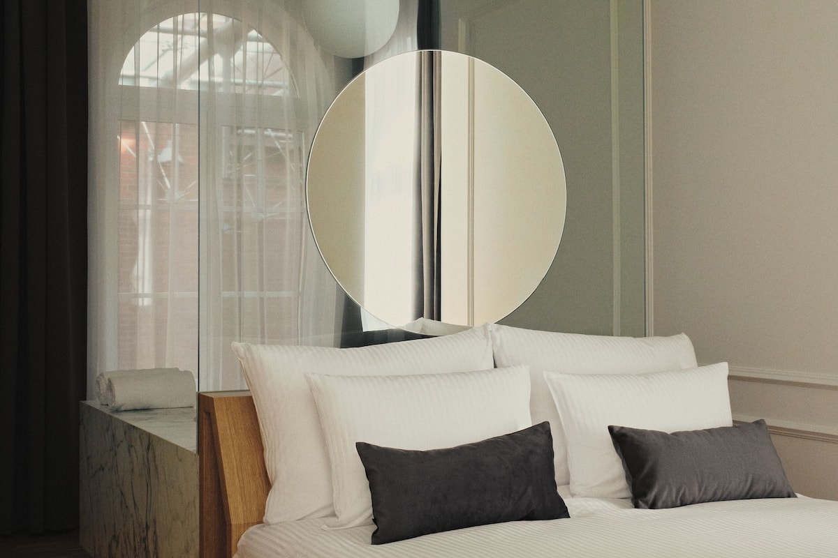 A modern bedroom with a large bed, a round mirror, and a view of a building through a window.