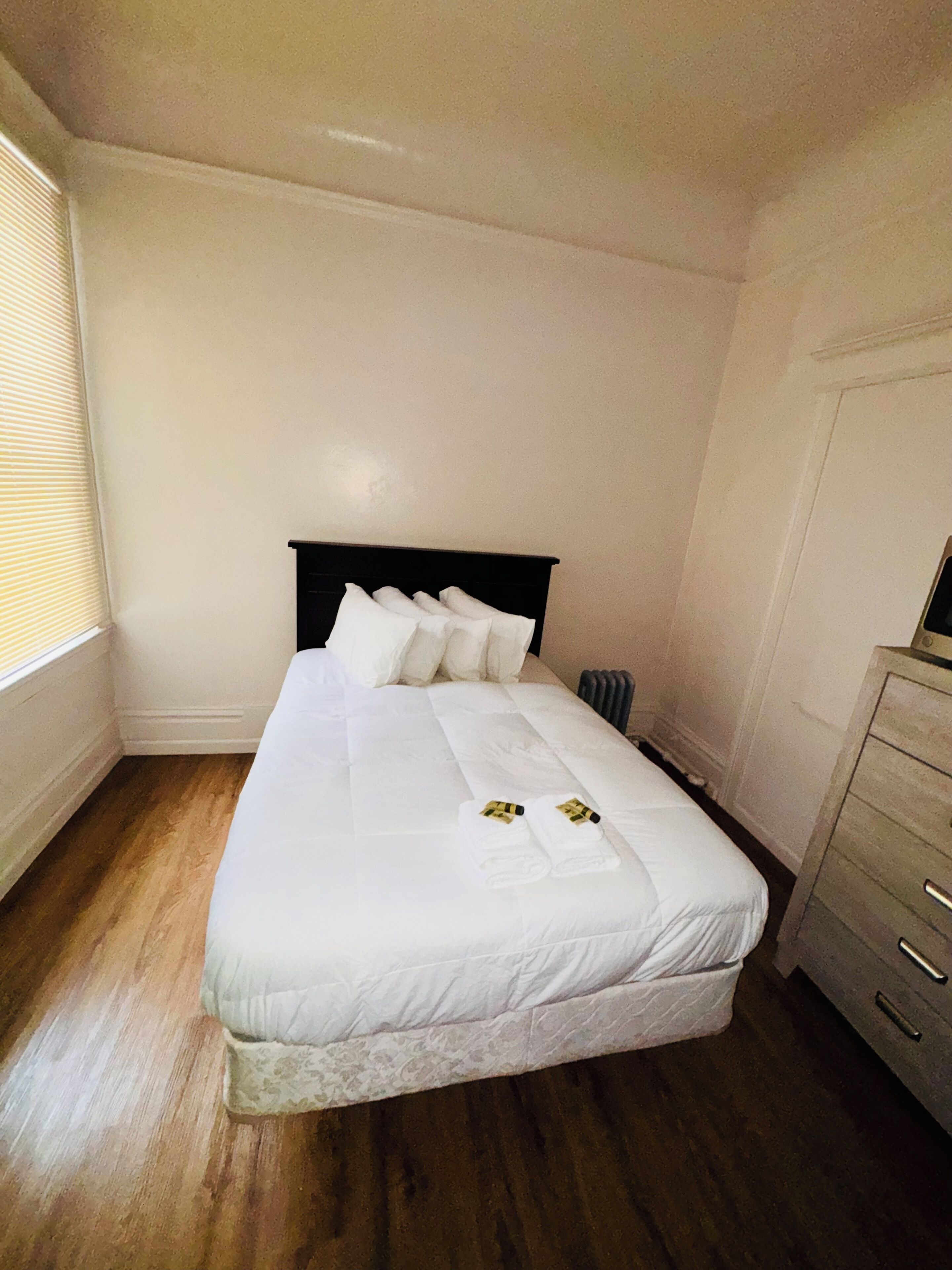 A neatly made bed with white linens and pillows, a wooden floor, and a radiator on the wall.