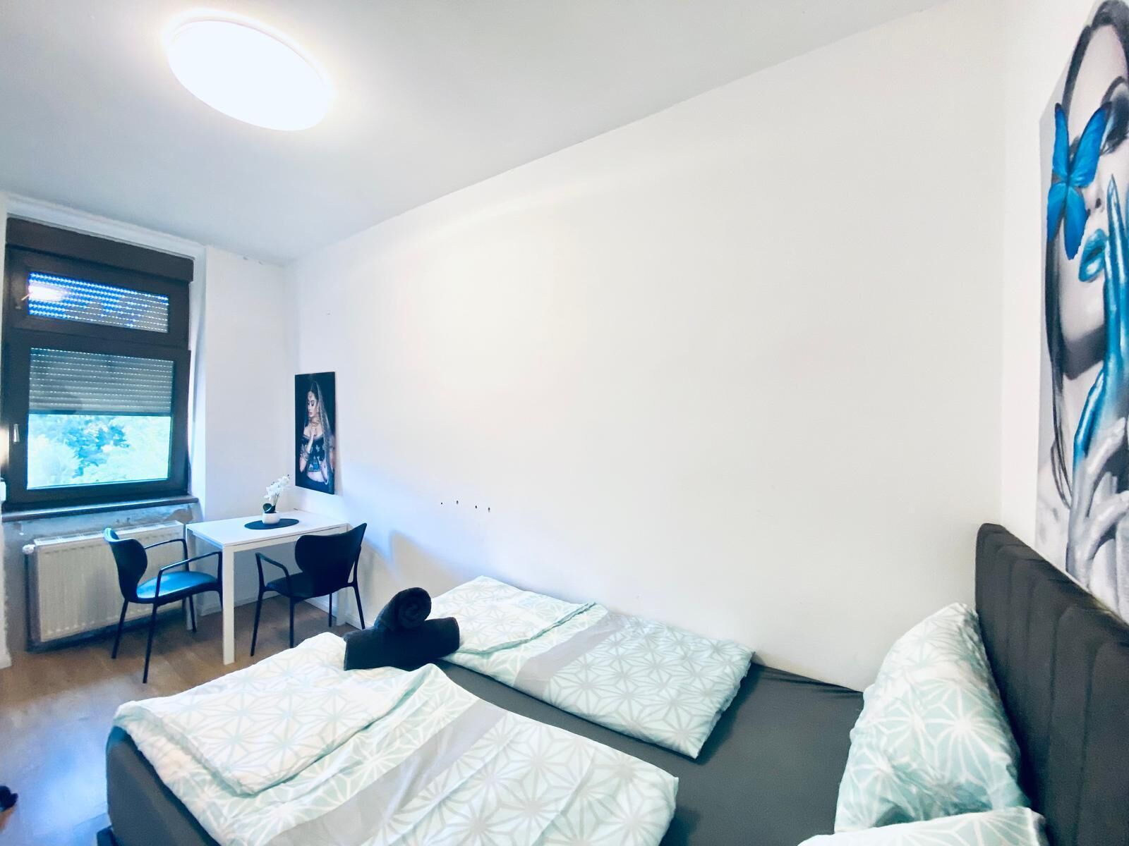 A small, clean room with a bed, a desk, and a chair. There is a window with blinds, a radiator, and a painting on the wall.