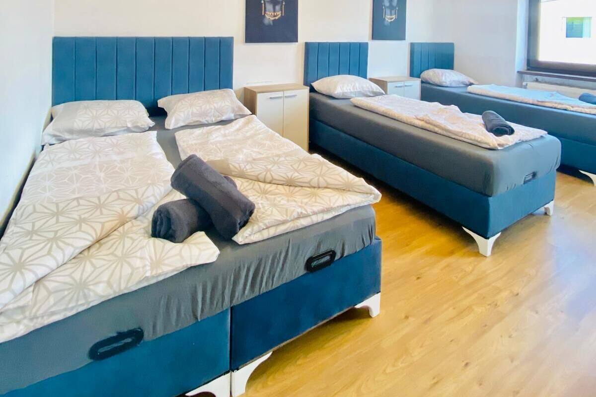 A hotel room with three beds, each with a blue headboard and white bedding. There are two wall-mounted artworks above the beds.