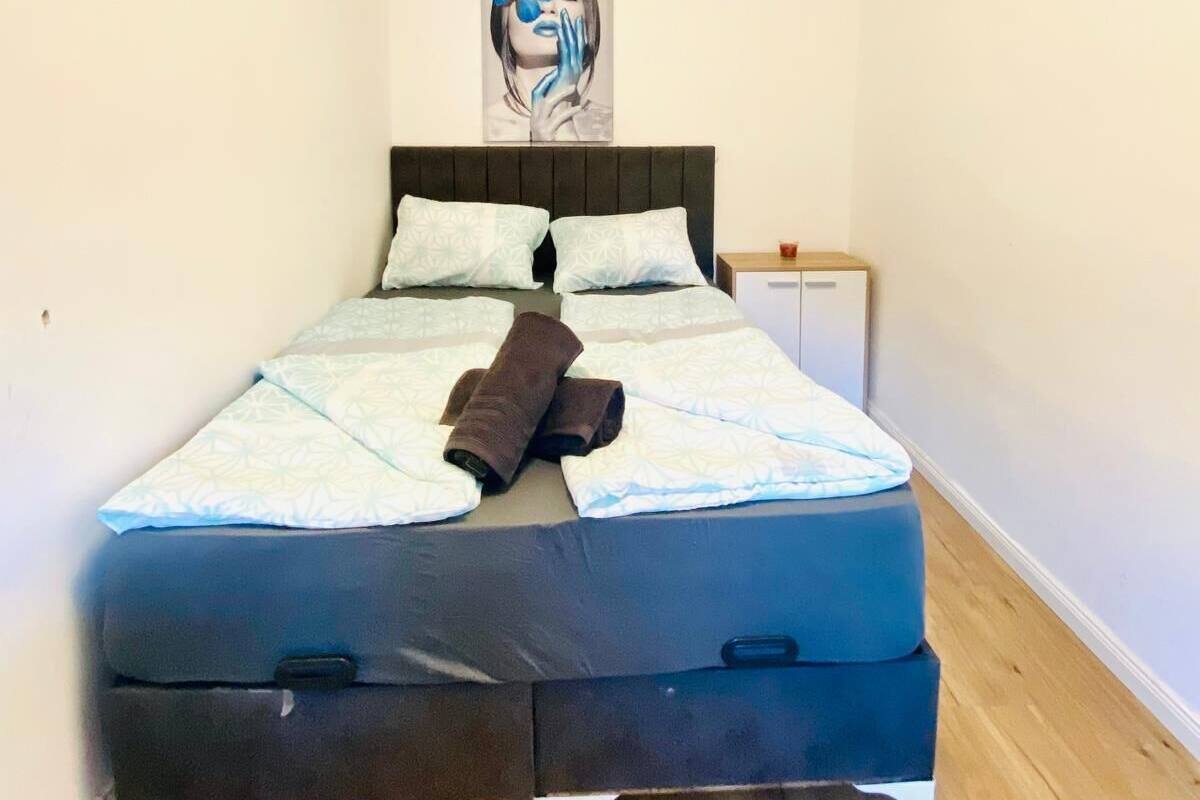 A neatly made bed with patterned bedding and a brown towel, a bedside table with a red object, and a painting on the wall.