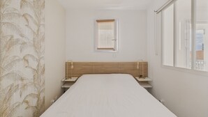 Una habitación de hotel estrecha con una cama, un banco de madera, una mesita pequeña, una lámpara de pared y una ventana con persianas.