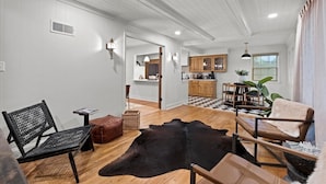 A spacious living area with a wooden floor, a black leather sofa, a brown leather chair, and a black leather rug. There is a kitchen area with wooden cabinets and a checkered floor.