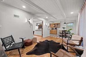 A spacious living area with a wooden floor, a black leather sofa, a brown leather chair, and a black leather rug. There is a kitchen area with wooden cabinets and a checkered floor. - The Glenwood - A Cozy Ranch in Mission, KS (Mission)