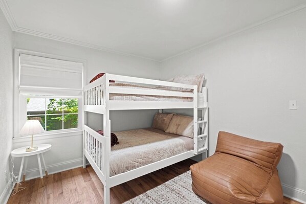 A bunk bed room with a window, a small table with a lamp, and a brown chair. - The Glenwood - A Cozy Ranch in Mission, KS (Mission)