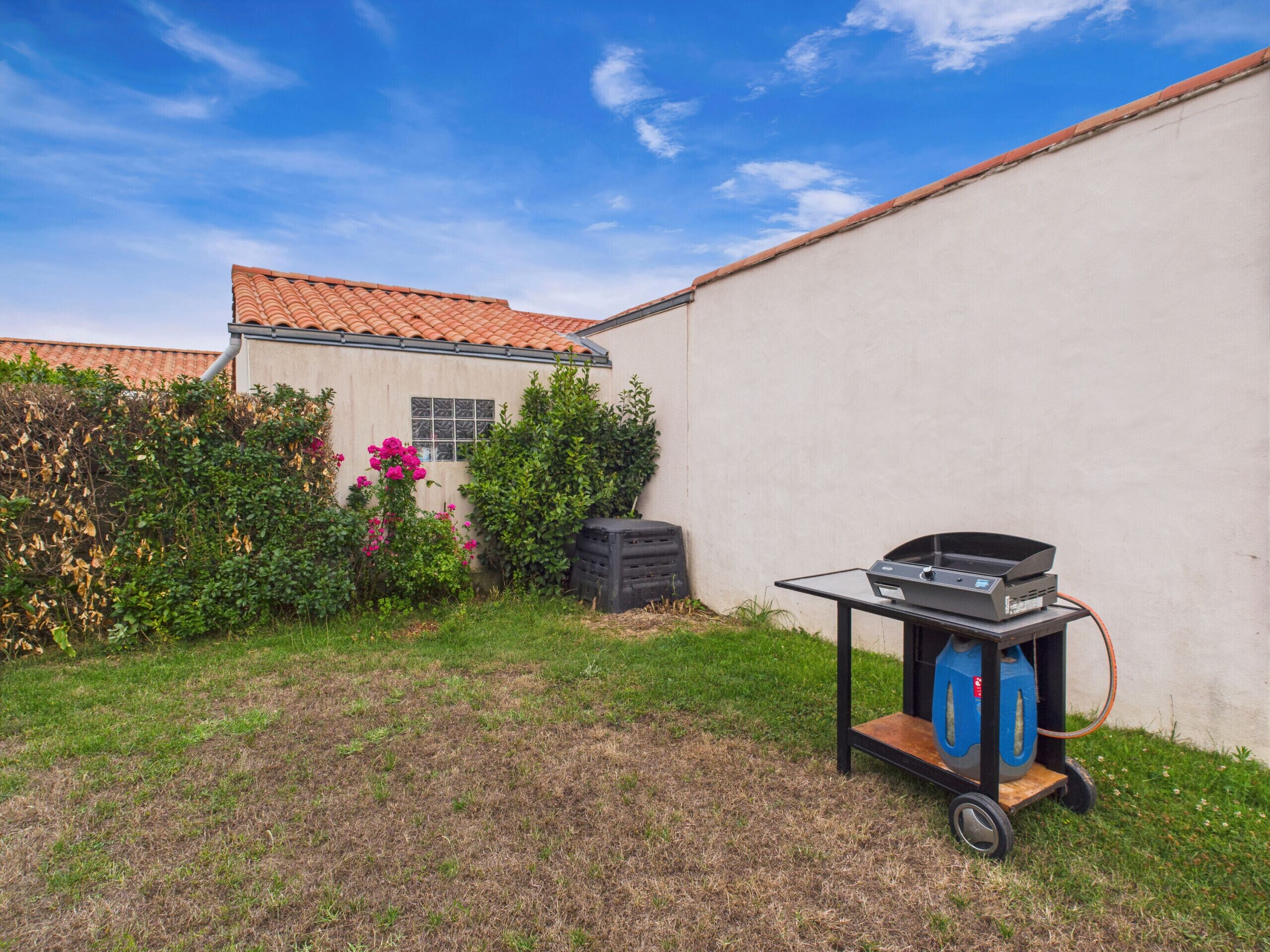 Un jardin avec un barbecue sur roulettes, un mur blanc et une maison avec un toit en tuiles.