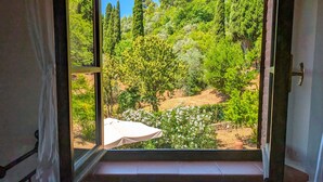 A room with a large window offering a view of a garden with trees and a white table. - Villa Poggio al Coniglio at Suvereto with Pool (Suvereto)