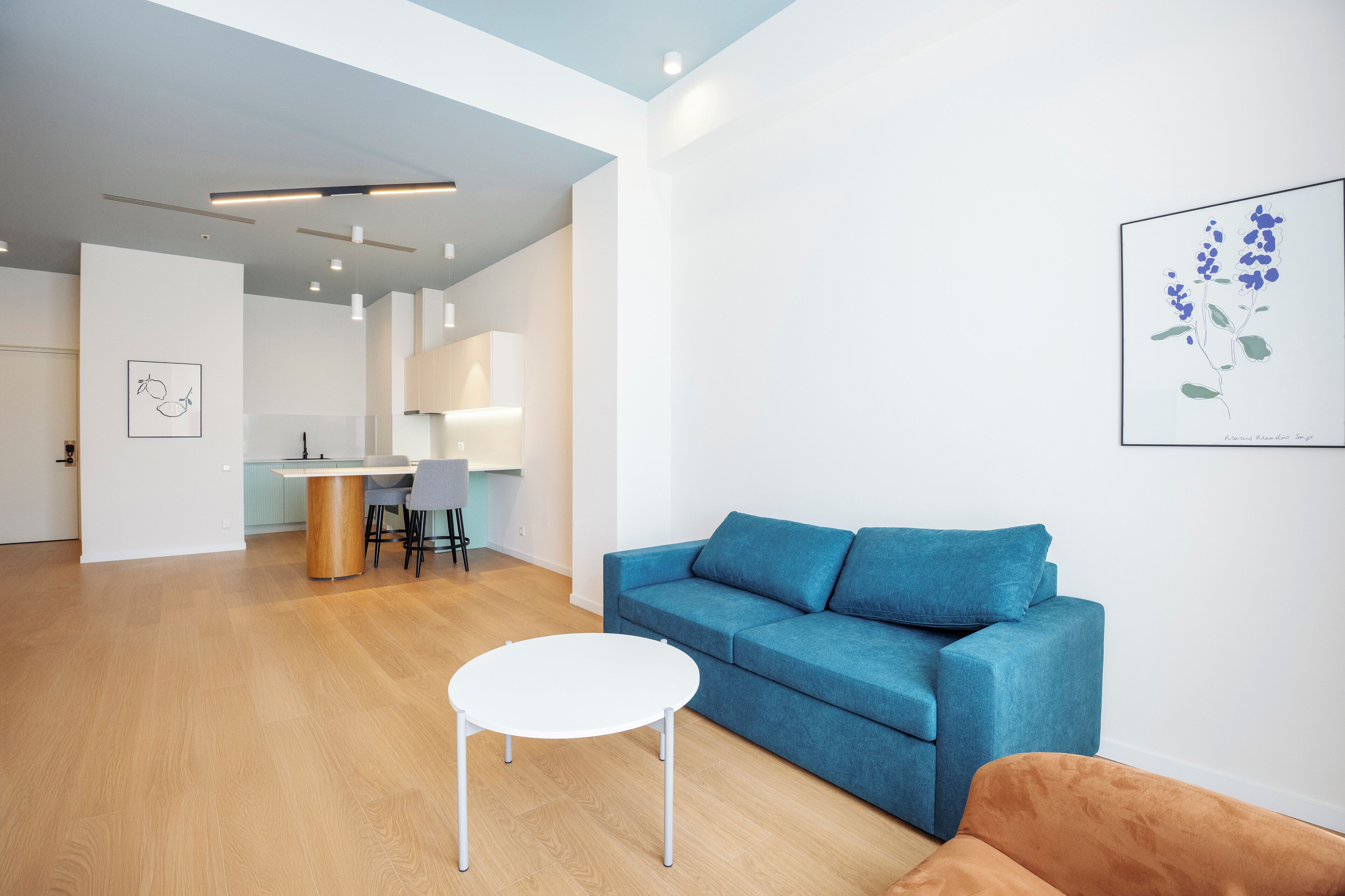 A modern living room with a blue sofa, a white coffee table, and a brown armchair. There is a kitchen area in the background with a dining table and chairs.