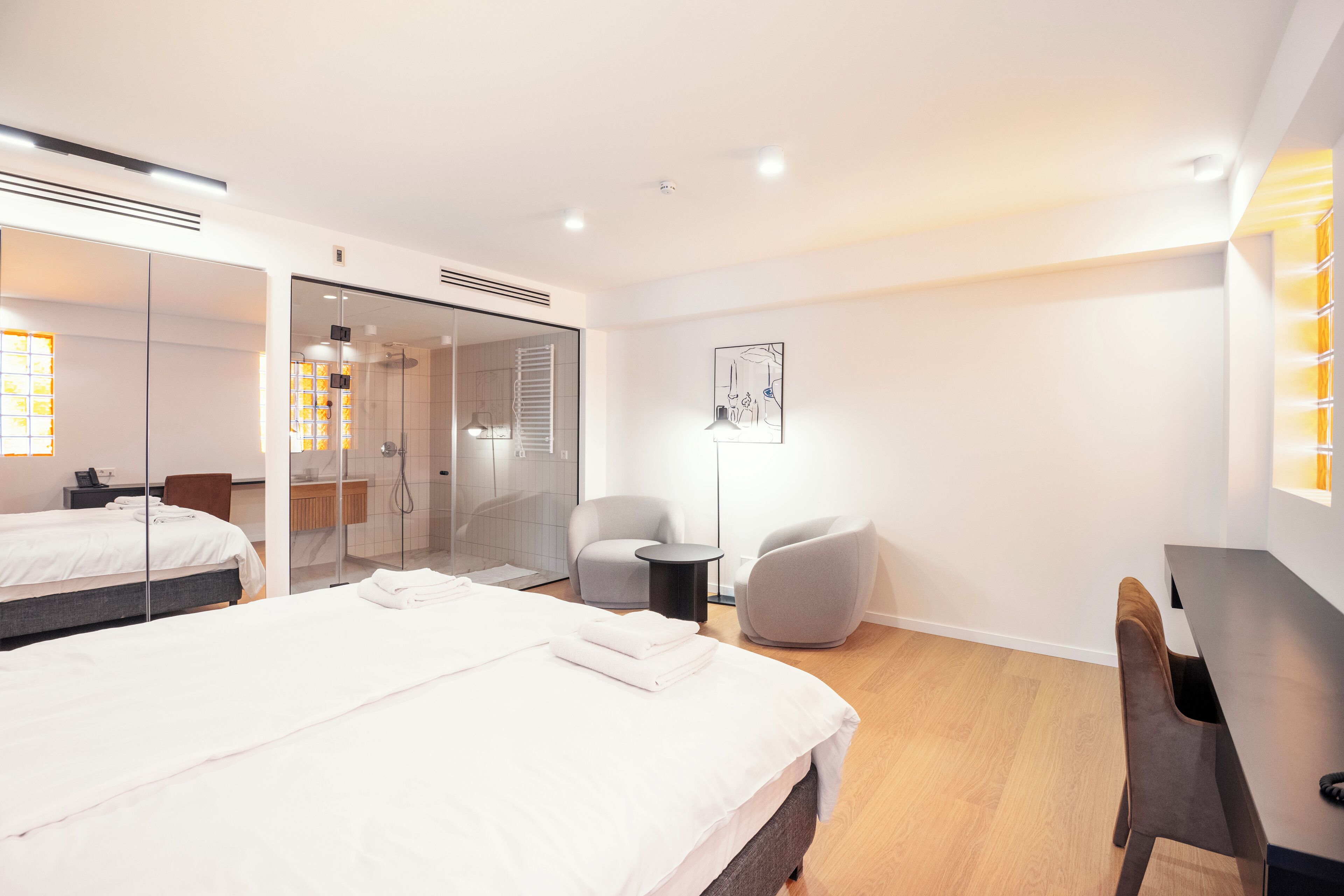 A modern hotel room with a large bed, a desk, a chair, a small table, a mirror, and a shower area.