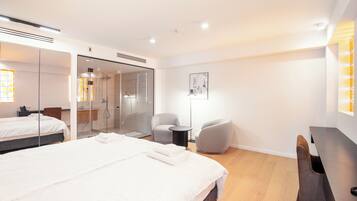 A modern hotel room with a large bed, a desk, a chair, a small table, a mirror, and a shower area.