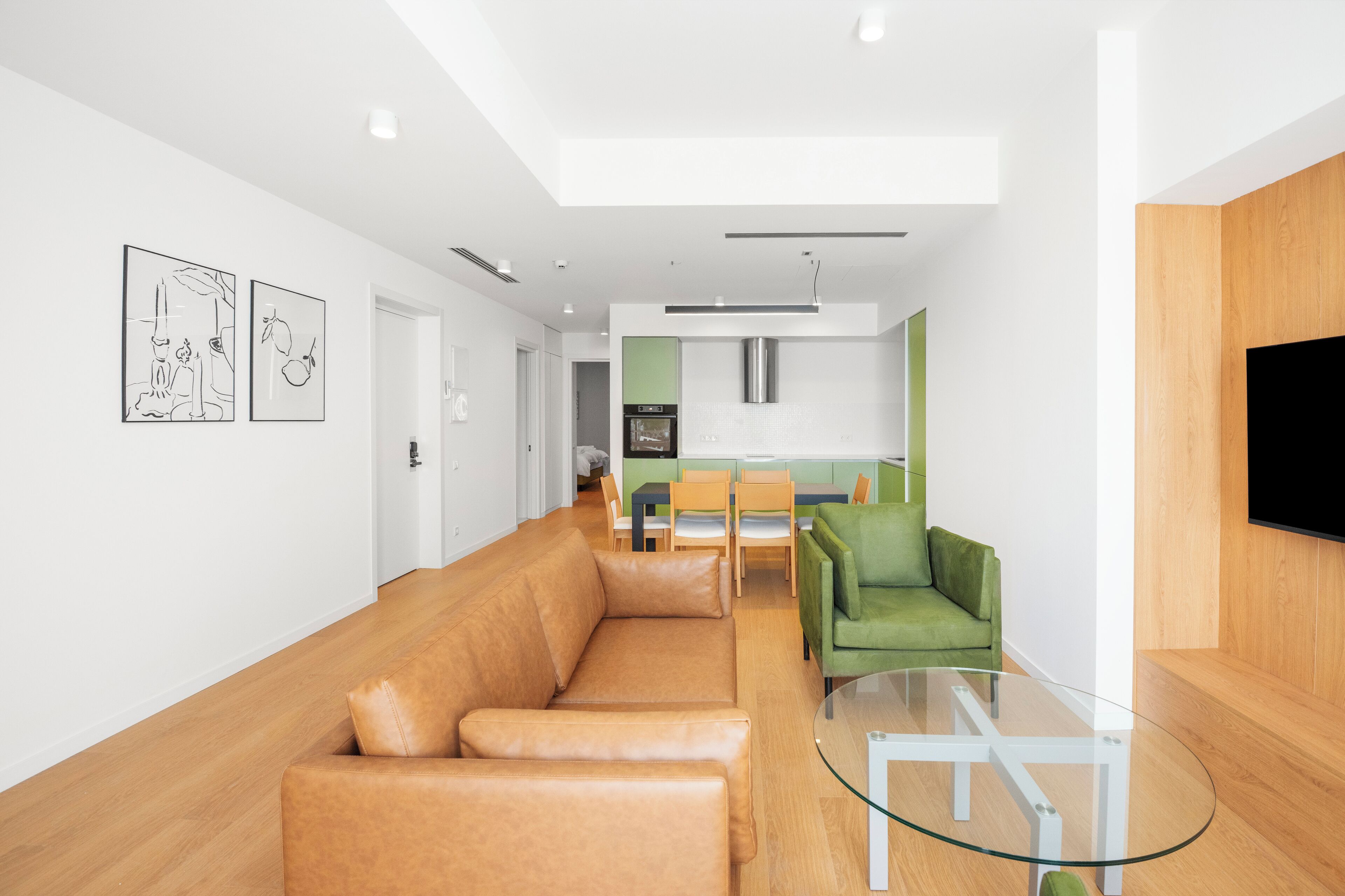 Una sala moderna con un sofá de cuero, una mesa de centro de vidrio y un sillón verde. Se ve un área de cocina al fondo con una mesa de comedor y sillas.