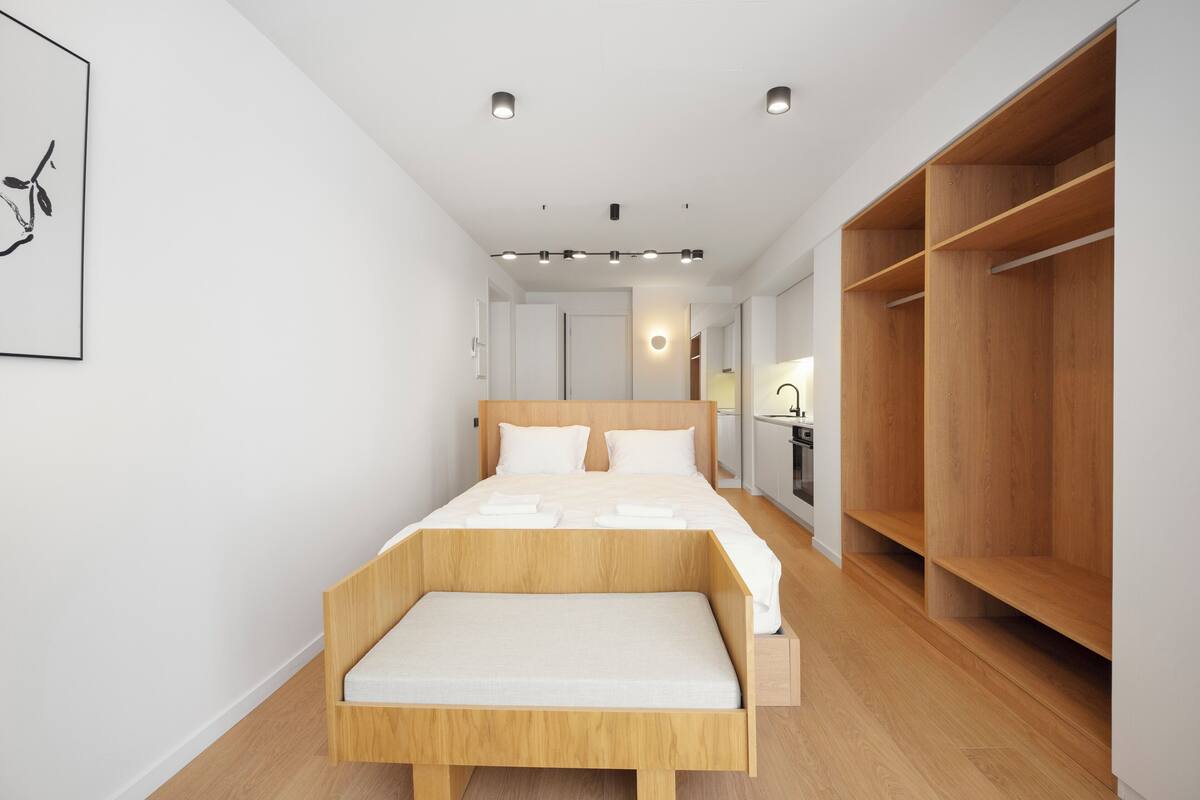 Een moderne slaapkamer met een houten bed, een bankje aan het voeteneind, een kledingkast en een kleine kitchenette.