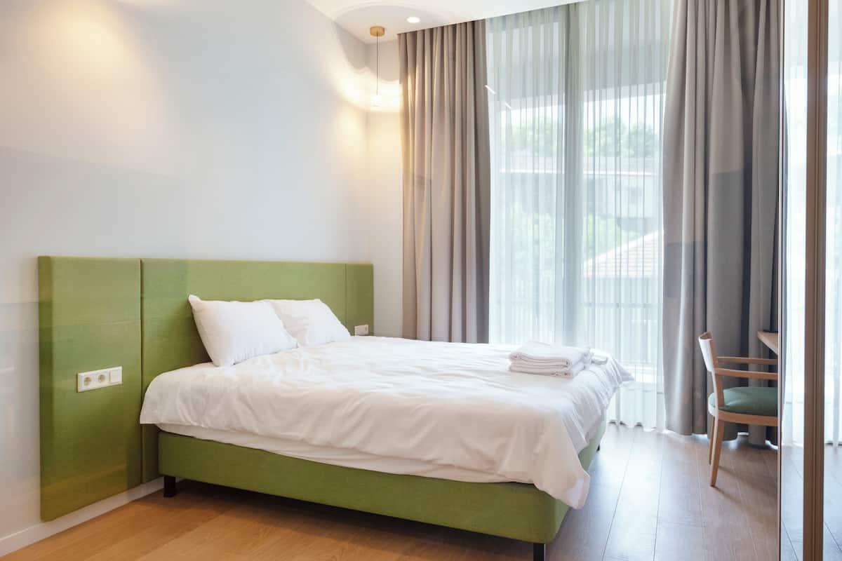 Een keurig opgemaakt bed met witte lakens, een groen hoofdbord en een houten stoel ernaast.