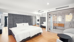 A modern hotel room with a large bed, a seating area, and a glass partition separating the bedroom from the bathroom.