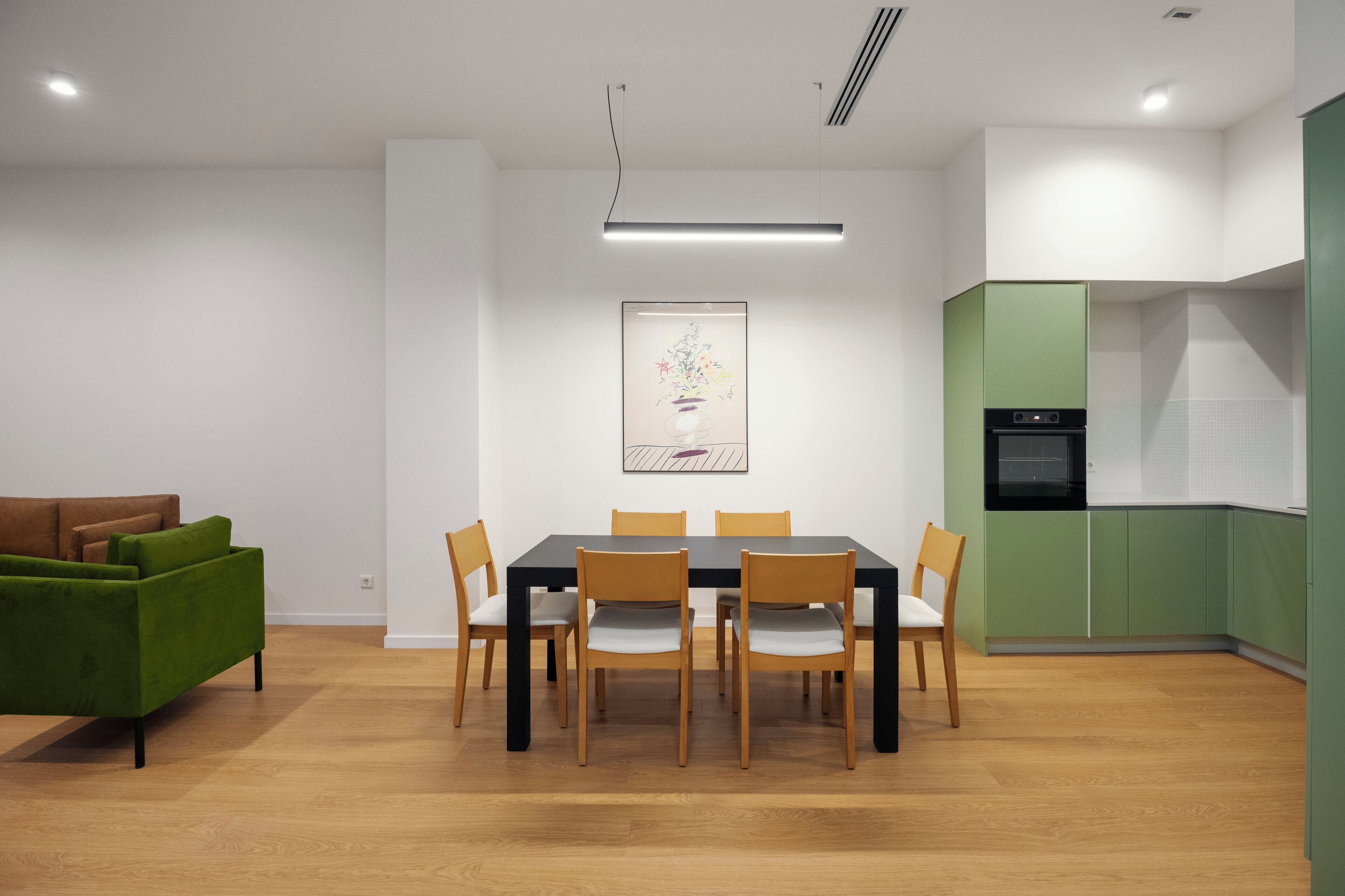 A modern dining area with a rectangular table and six chairs, a green sofa, and a kitchen with green cabinets and a black oven.