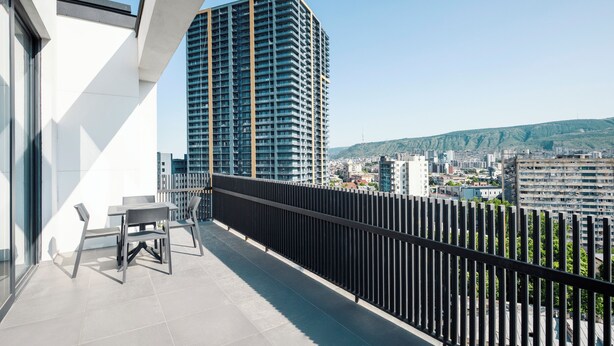 A balcony with a view of a tall building, a table with chairs, and a black railing.
