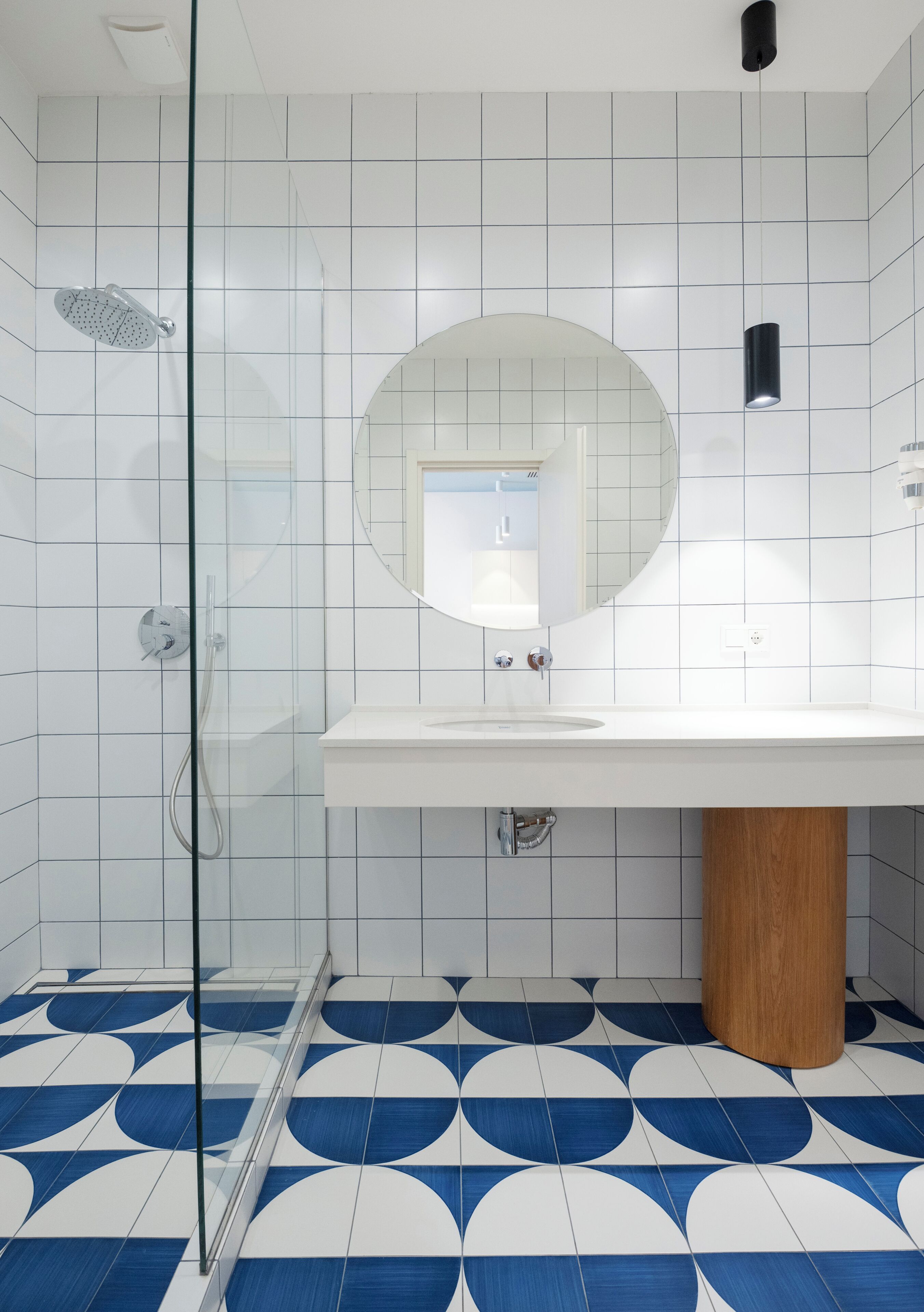 A modern bathroom with a glass shower enclosure, a round mirror above a white sink, and a patterned floor.
