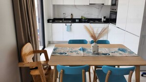 Una cocina moderna con armarios blancos, encimera negra y electrodomésticos de acero inoxidable. Hay una mesa de comedor con sillas azules y un jarrón con plantas secas.