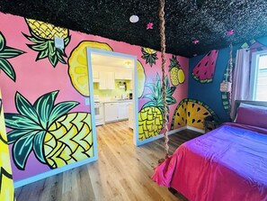 Una habitación con un mural llamativo de piña, una cama con colcha rosa y una cocina visible a través de una puerta.