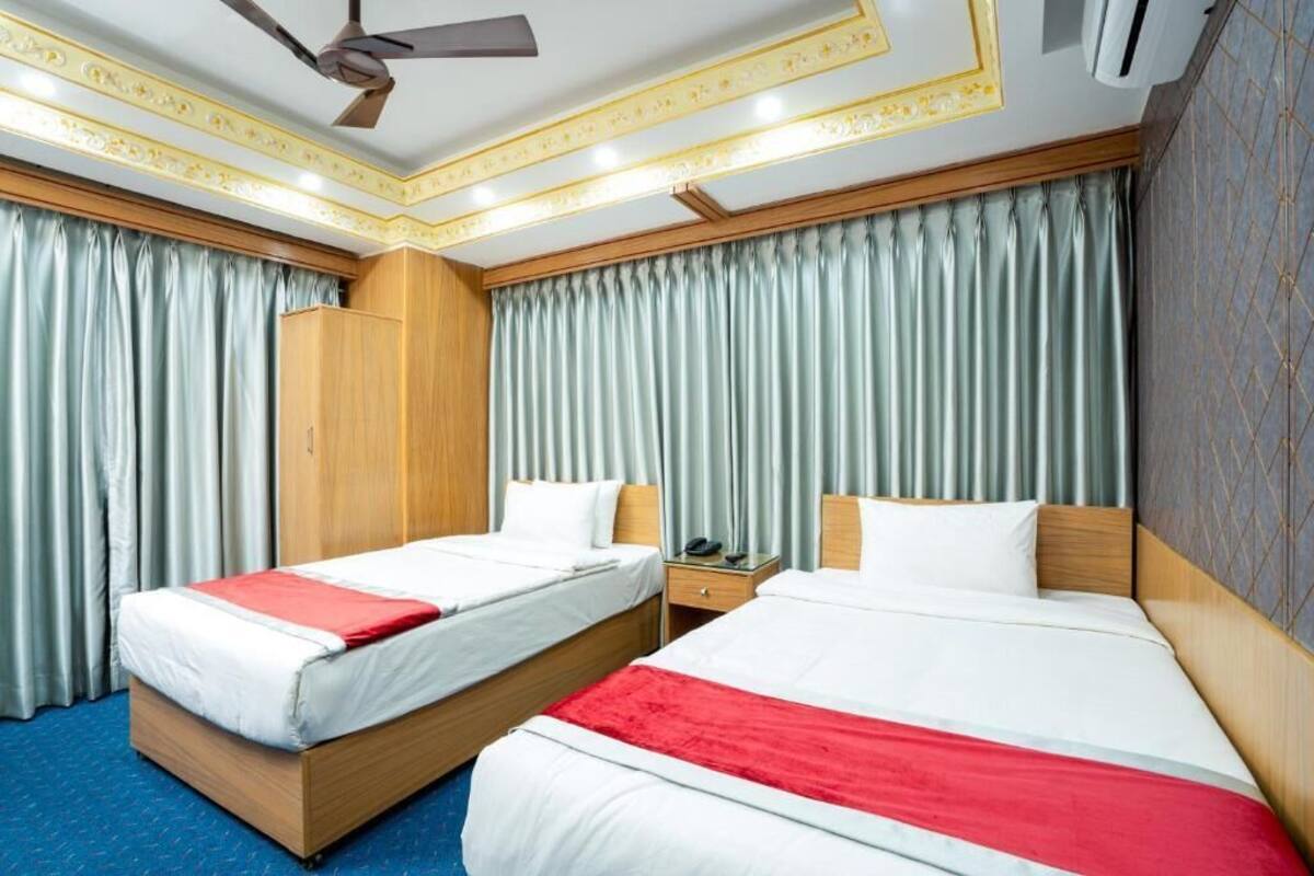 A hotel room with two beds, a wooden wardrobe, a ceiling fan, and a large window with curtains.