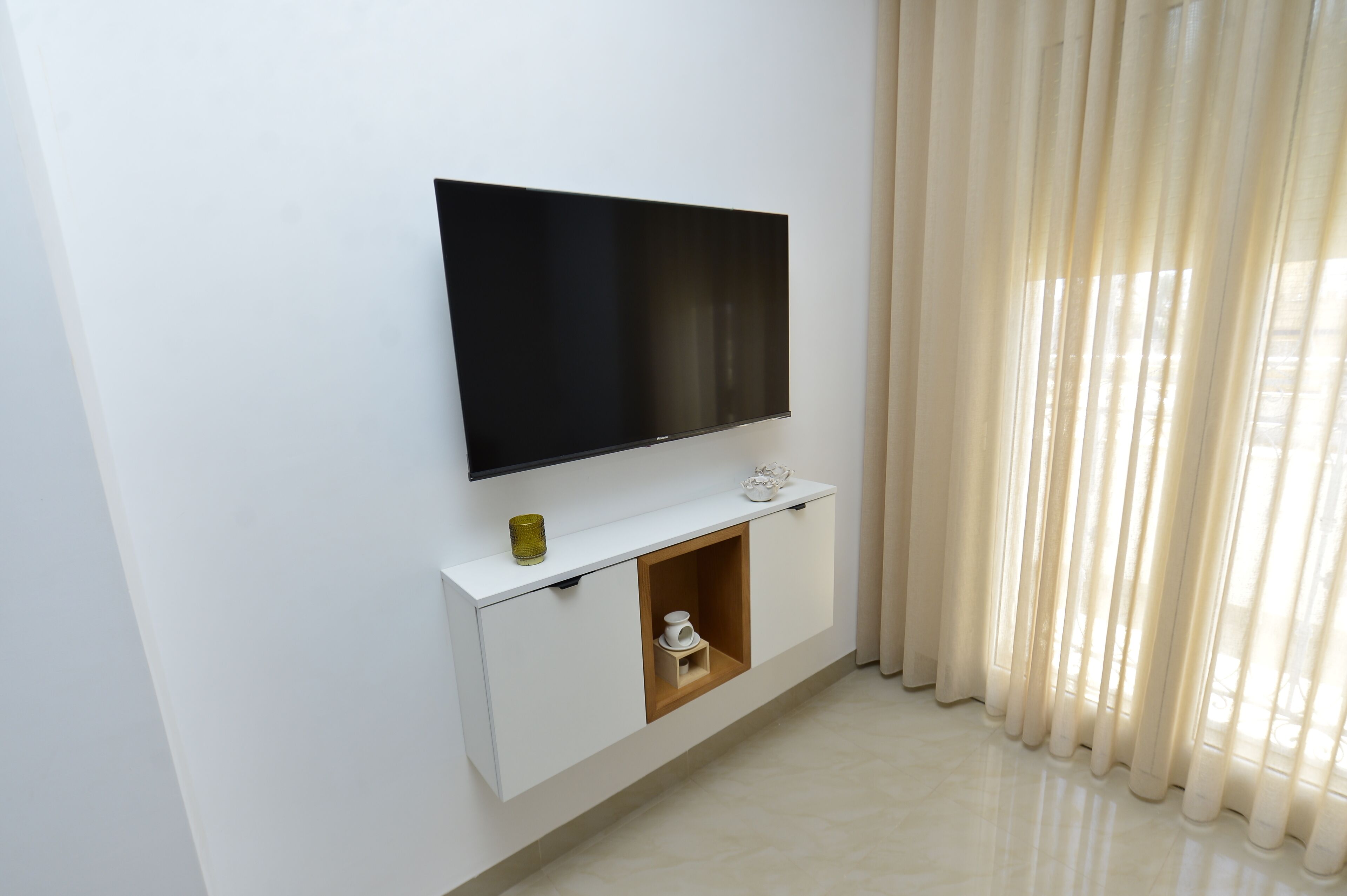 A modern living room with a flat-screen TV mounted on a white cabinet, a small shelf with decorative items, and a wooden cabinet with a cup.