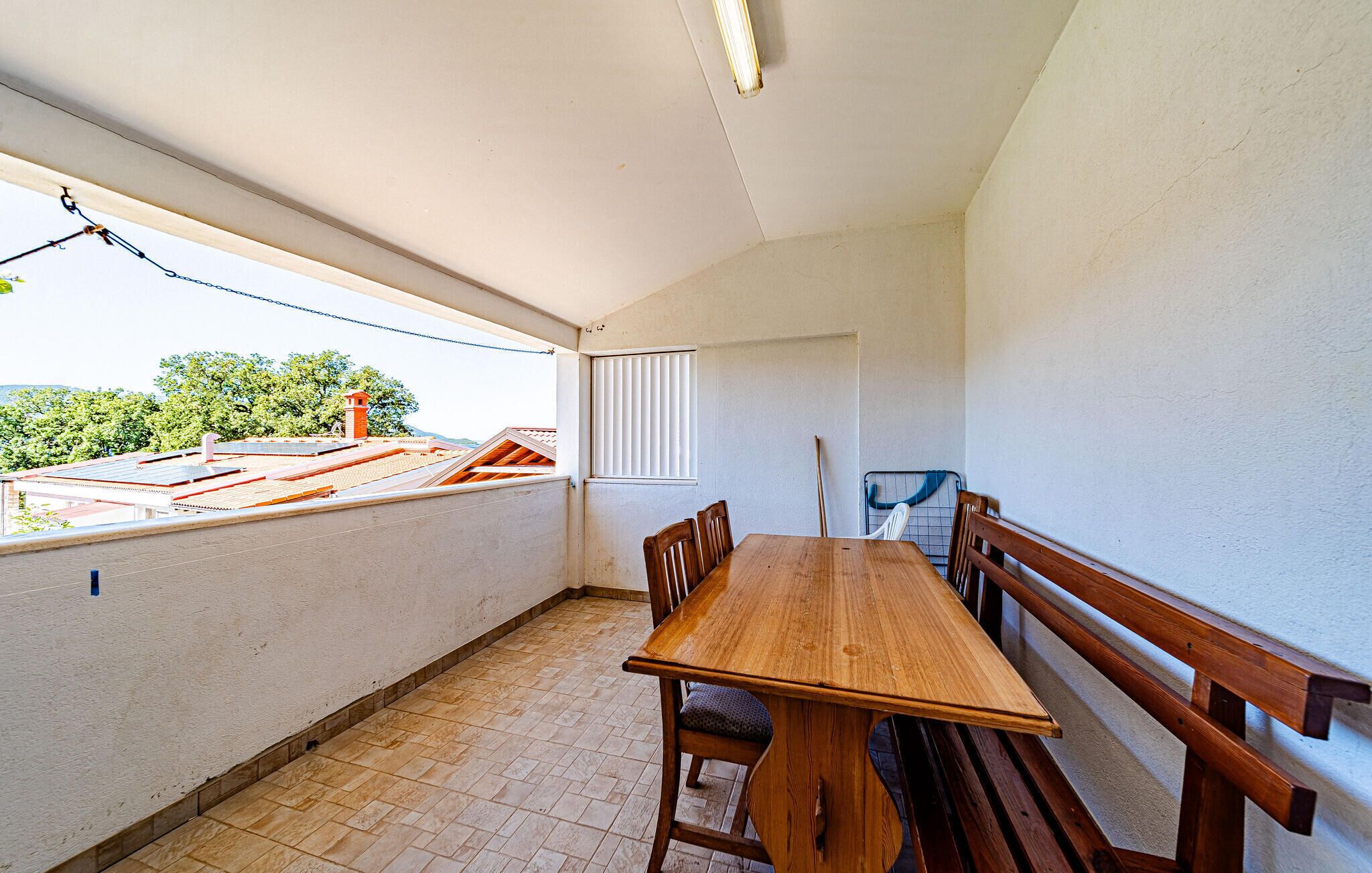 Un balcón con mesa y bancos de madera, una ventana con persianas y vistas a tejados y árboles.