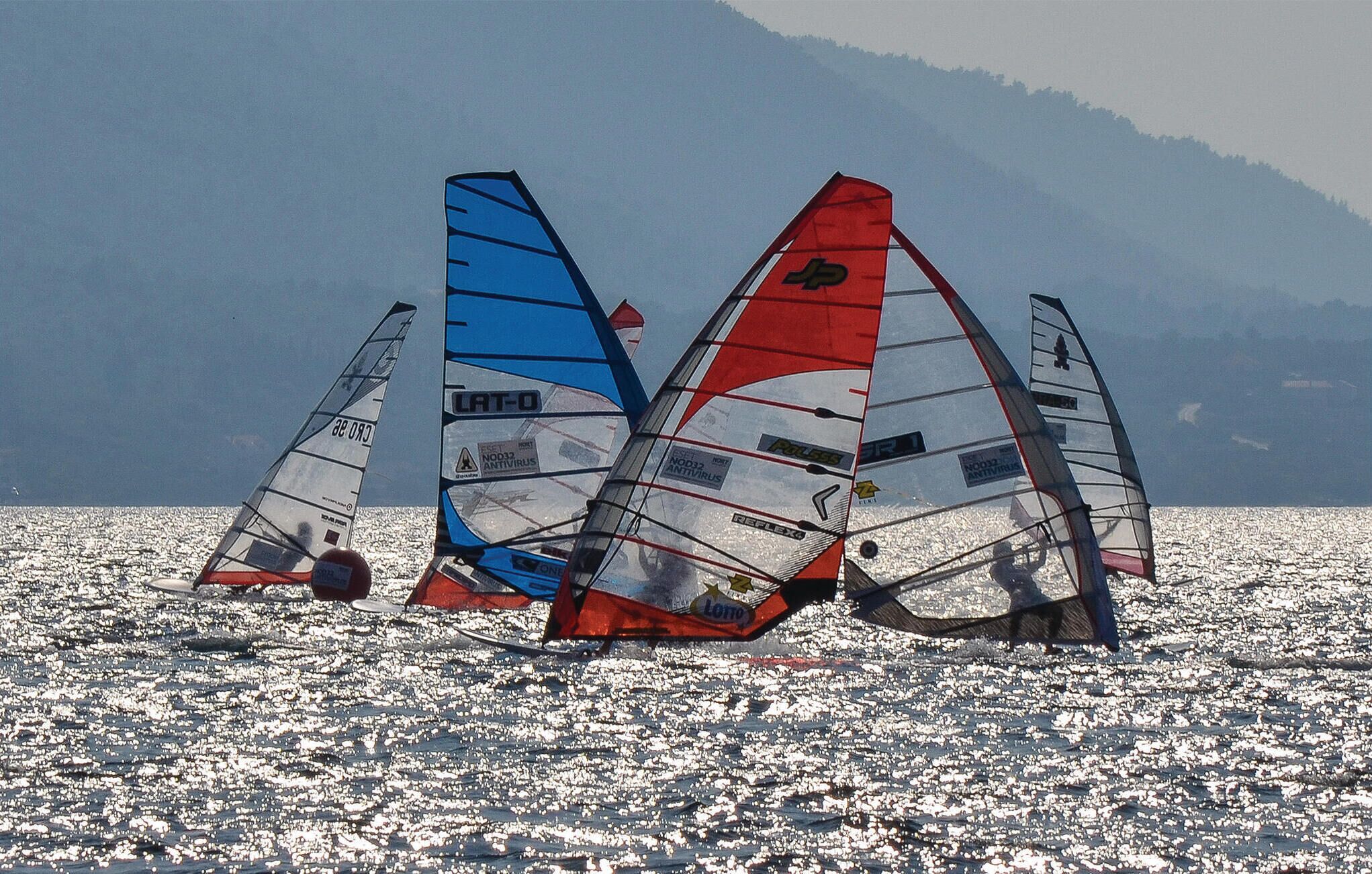 Tablas de windsurf sobre una superficie acuática reflectante, con montañas al fondo.